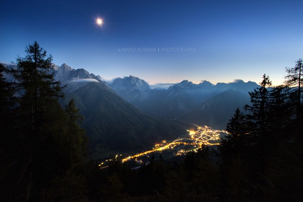 The city lights down in the Upper Sava valley illuminate streets in Kranjska Gora area. The moon is high up over the Julian Alps mountains. The walls of the highest peaks are still illuminated by the weak western skylight and moonlight. 